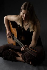 Photo of Kate Weekes seated cross-legged playing guitar