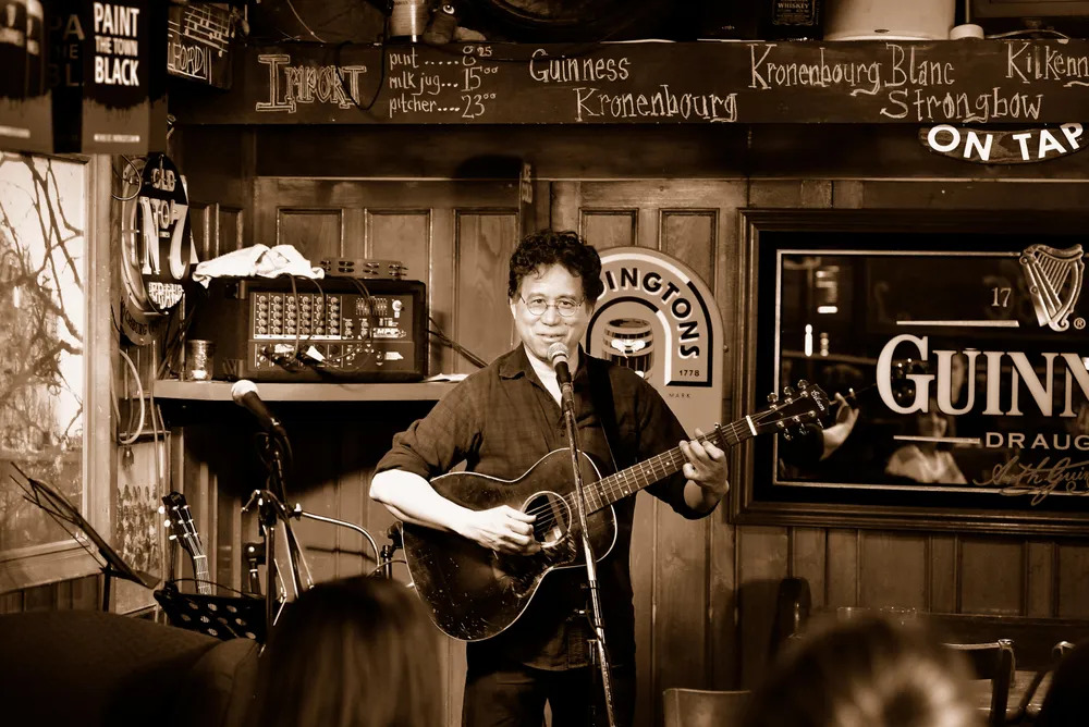 Photo of Vince Halfhide playing his guitar at a microphone, in the corner of a bar
