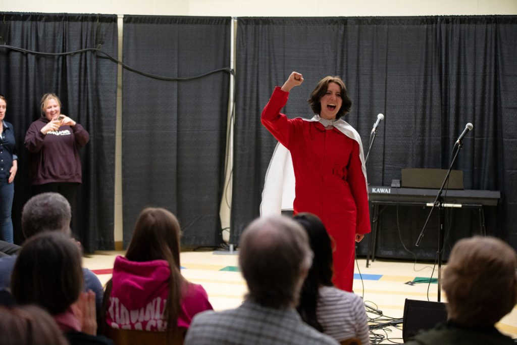 A performance by The Algonquin College Emerging Stars, wearing red coveralls and a white cape, raising her fist and calling out