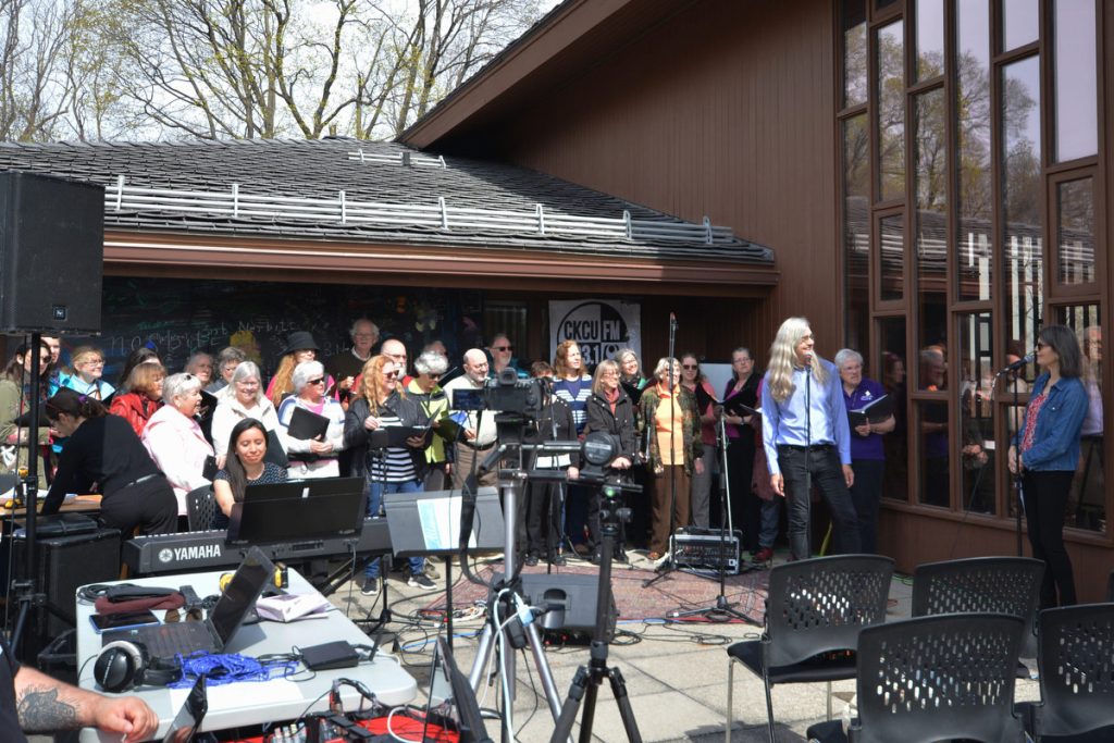 A chorus of voices outdoors singing for CKCU's broadcast, on the upper deck at First Unitarian Church