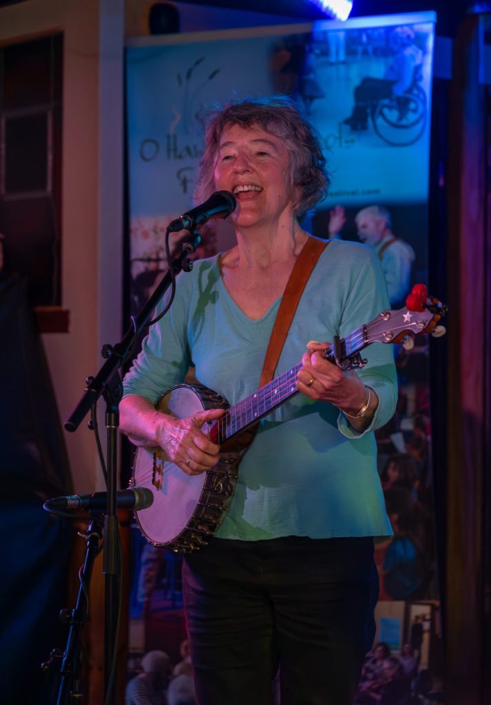 Mary Gick playing and singing at Irene's Pub, in front of a festival banner and smiling away!
