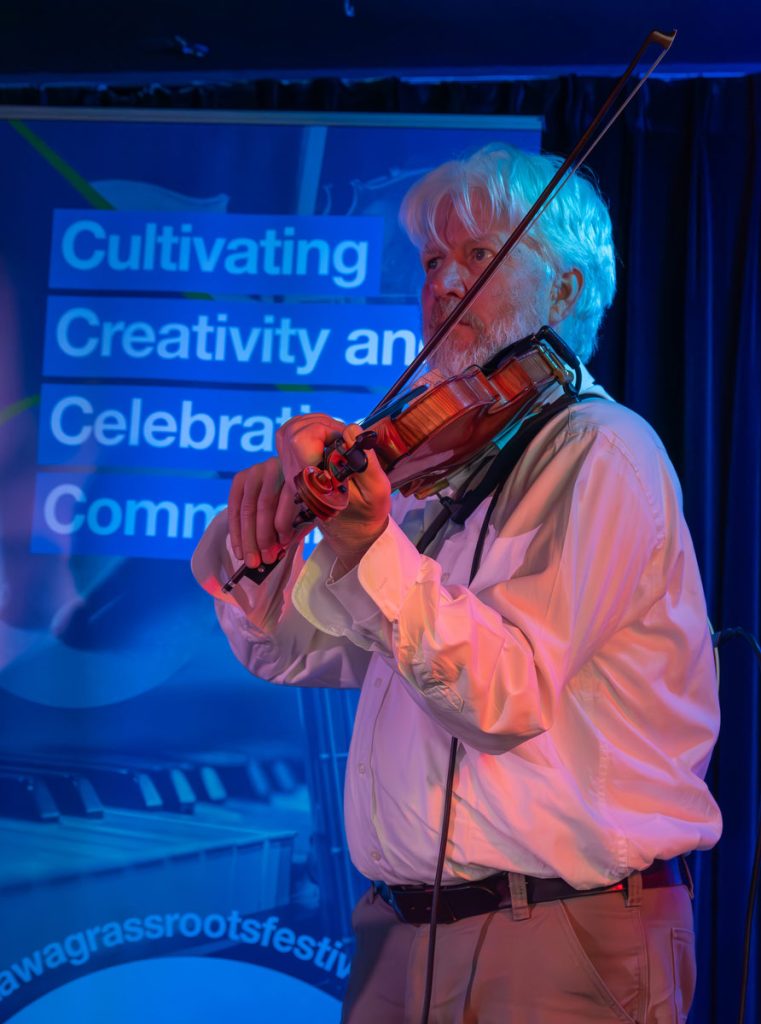Michael Ball plays fiddle on stage at Irene's Pub in front of a festival banner that says "Cultivating Creativity and Celebrating Community"